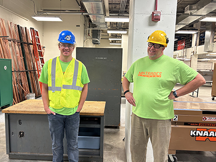 Jack Roessler and fellow MTA Trainer stand in a woodshop wearing construction gear